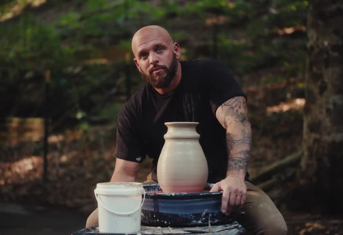 Portrait of a serious, tattooed potter sitting with his finished clay vase on a wheel in a quiet forest setting