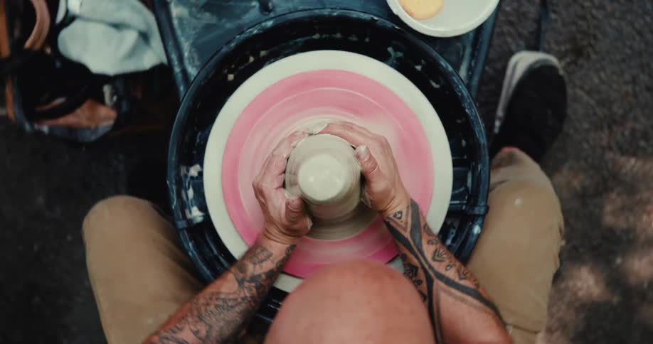 Overhead shot of a bald man with tattoos shaping a piece of clay on a spinning potter's wheel