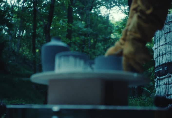 A male artisan with tattoos carefully places his handmade ceramic pottery on a stand in a serene forest setting