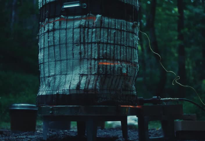 A homemade raku kiln glows red-hot from the intense firing process in a dark forest setting