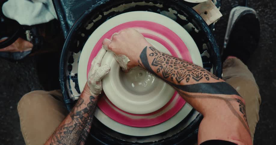 Overhead view of a tattooed artist skillfully shaping a lump of clay on a spinning pottery wheel