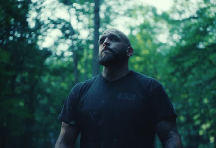 A rugged, muscular man with tattoos stands alert in a dense, green forest