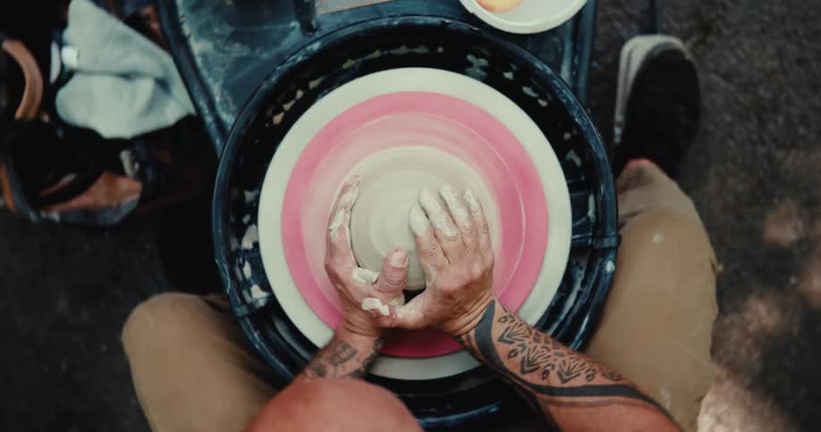 A top-down view of a tattooed artist expertly shaping a lump of clay on a spinning pottery wheel