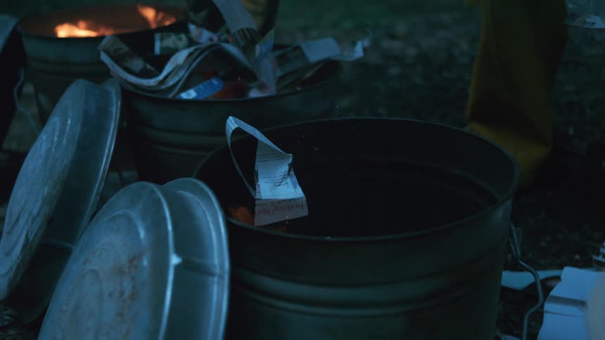 Flames slowly consume pieces of paper dropped into a metal burn barrel outdoors at night