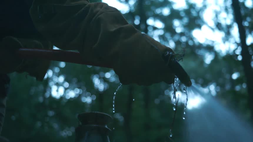 A firefighter's gloved hand holds a spraying fire hose nozzle in slow motion against a dark forest background