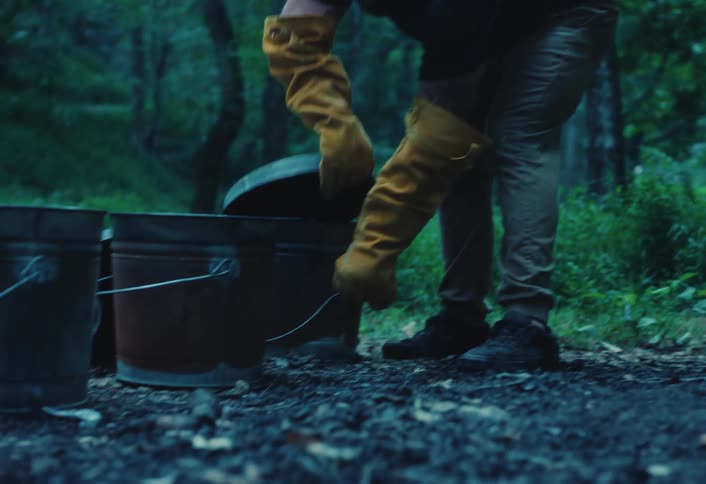 A craftsman in protective yellow gloves tends to metal reduction chambers during the Raku pottery firing process in a forest setting
