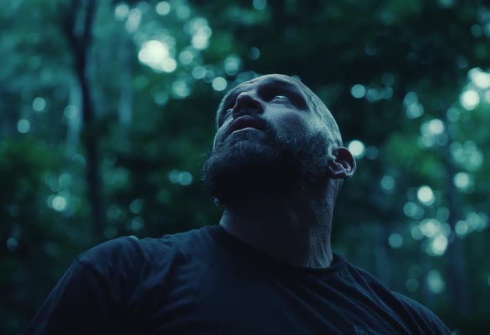 A tired, bearded man in work gloves takes a break in a dark forest, looking up before wiping his brow
