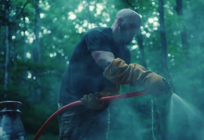 A man in protective gloves sprays steaming hot containers with a water hose in a misty forest setting