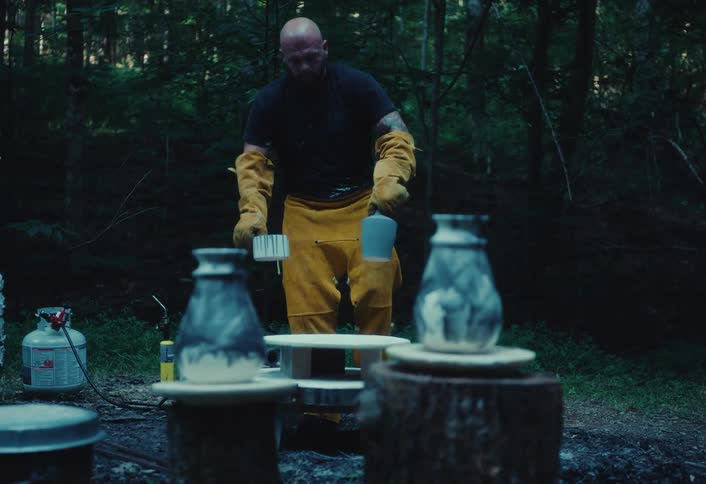 A focused craftsman in protective gear carefully prepares his handmade pottery for an outdoor firing in a rustic, wooded setting