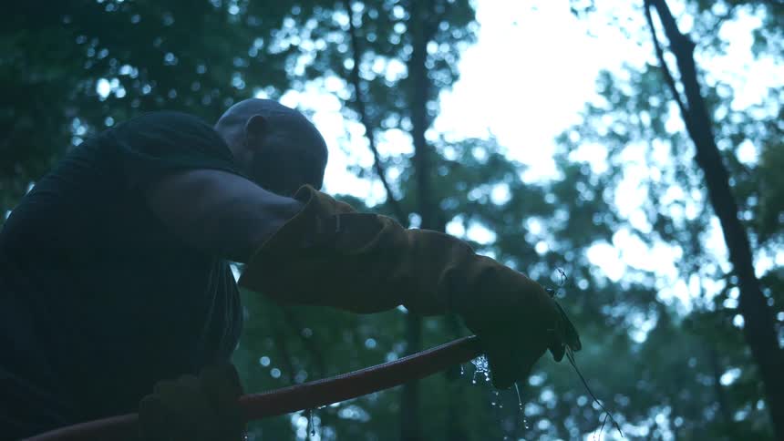 Low angle slow motion shot of a silhouetted worker in heavy gloves handling a leaking red water hose in the woods