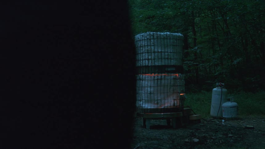 A handmade propane-fueled kiln glows with an orange fire in a dark, remote forest setting at dusk