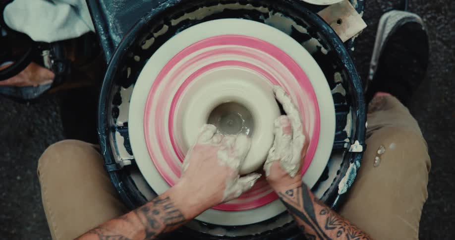 A top-down view of a tattooed artist skillfully shaping a clay pot on a spinning pottery wheel