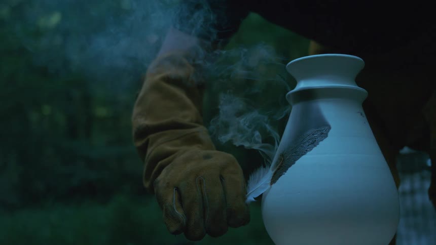 An artist carefully burns a feather onto a hot ceramic vase, leaving a smoky design on the surface in slow motion
