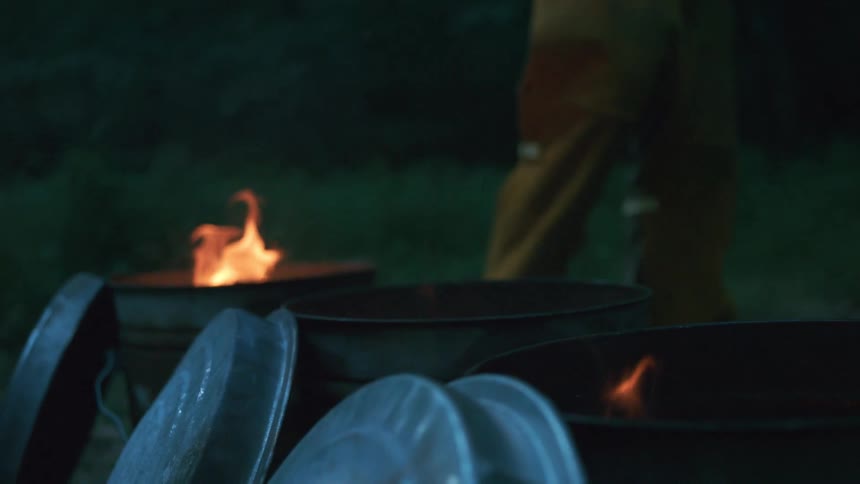 Slow-motion close-up of fires burning in metal barrels at night with a worker in the background