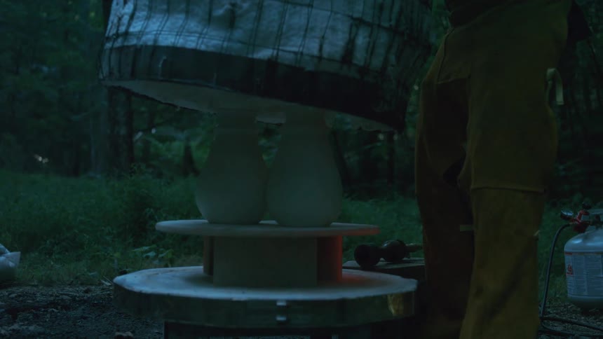 A potter wearing protective gear slowly lifts a hot raku kiln to reveal two glowing white vases in an outdoor setting at dusk