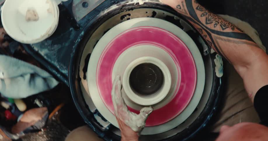 A top-down view of a tattooed man's hands skillfully shaping a clay pot on a spinning potter's wheel