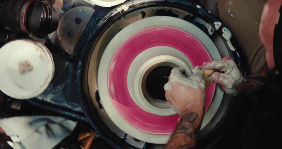 A top-down view of a tattooed artist's hands skillfully shaping a clay pot on a spinning pottery wheel