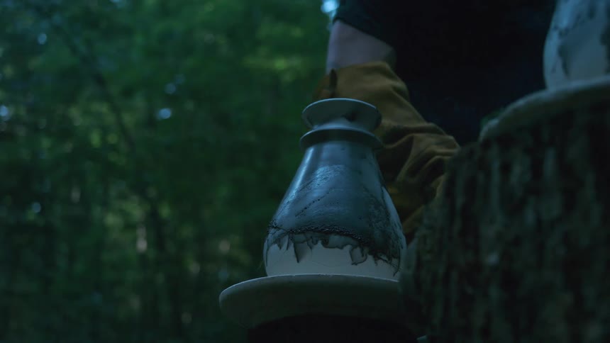 A steaming Raku-fired ceramic pot cools in a tranquil forest setting, with a gloved hand gently approaching
