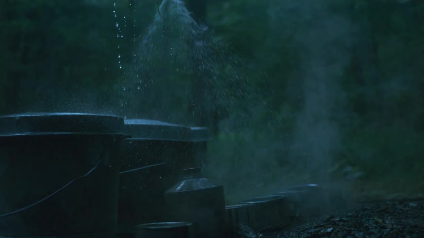 In slow motion, a gloved hand sprays misty water from a hose onto old metal containers in a dark, atmospheric forest