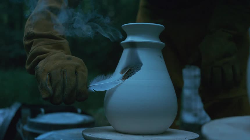 In a slow-motion close-up, an artisan carefully burns a feather onto a hot ceramic vase, leaving behind a smoky, dark pattern