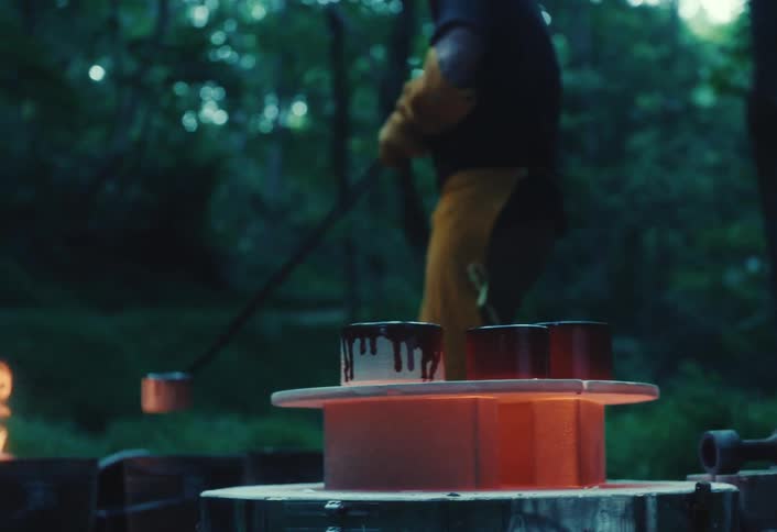 An artisan carefully removes a red-hot ceramic pot from an outdoor kiln during a Raku firing process in the woods