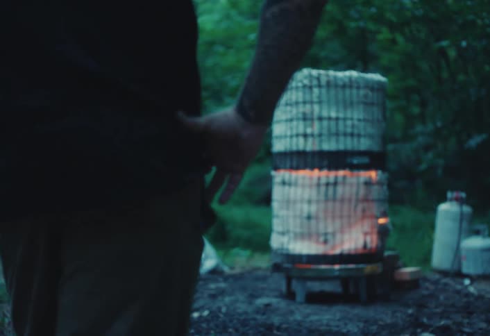 A man with tattooed arms watches his homemade raku kiln glowing with intense heat during an outdoor firing in the woods