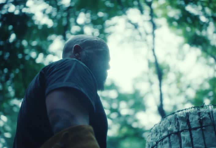 Low-angle shot of a muscular, bearded craftsman intently observing his handmade kiln in a dense, green forest