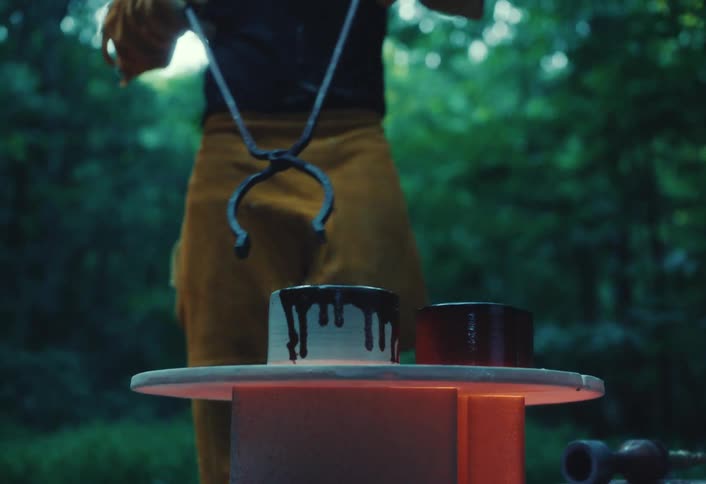 A potter carefully removes a red-hot glazed pot from an outdoor kiln with metal tongs in a lush forest setting