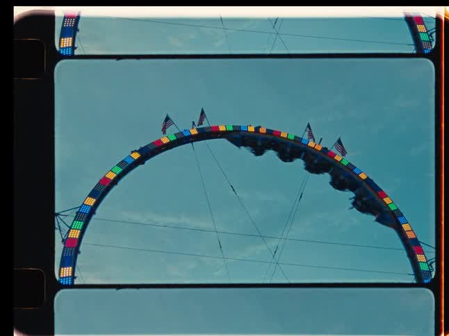 A vintage film-style shot captures people hanging upside down on a colorful carnival ride adorned with American flags against a pale blue sky