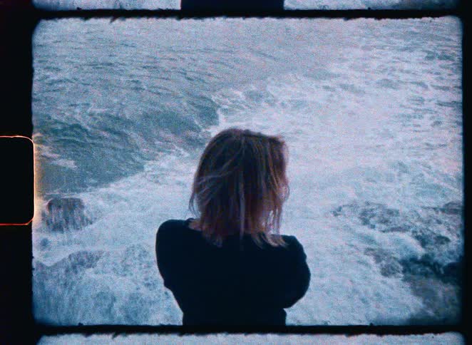 Vintage 8mm footage shows a solitary woman looking out at the powerful, churning ocean from a rocky coastline