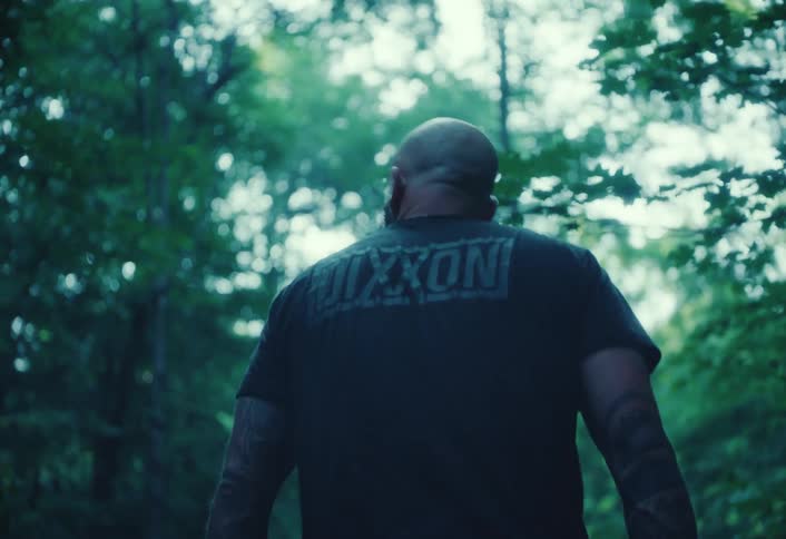 Slow motion shot following a muscular, tattooed man from behind as he walks down a grassy path in a dense, green forest