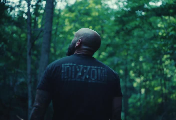 A bald, muscular man wearing a black shirt with tattoos walks slowly through a lush forest in slow motion