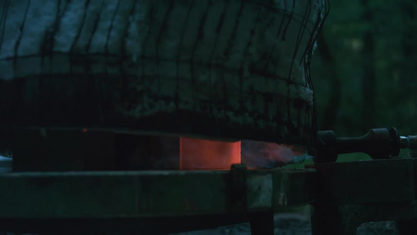 A close-up of an outdoor pottery kiln glowing red hot as flames and smoke escape during an evening firing