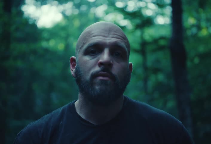 Close-up portrait of a serious, bald man with a beard staring intently into the camera in a moody forest setting