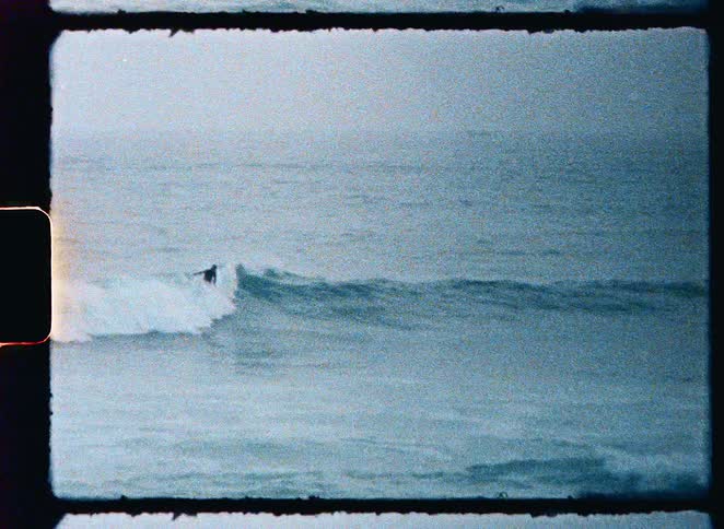 Slow-motion vintage 8mm footage shows a lone surfer riding a wave in the ocean on an overcast day