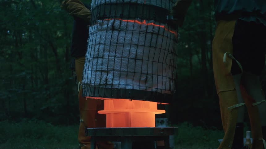 Artisans in protective gear slowly lift a homemade kiln, revealing a glowing red-hot ceramic sculpture at dusk