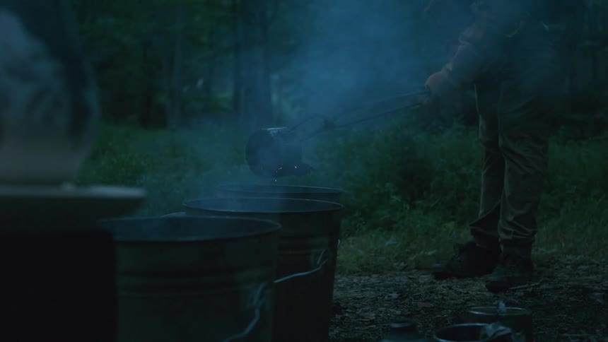A person uses tongs to remove a glowing hot object from a smoking bucket during a rustic firing process at dusk