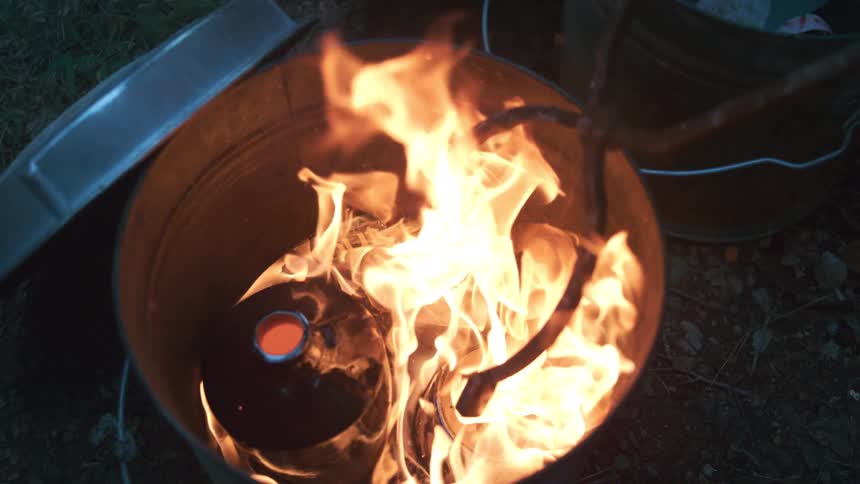 Vibrant orange flames dance in slow motion inside a metal burn barrel at dusk