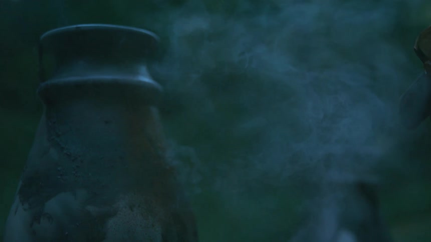 A mysterious cloud of smoke billows out of an old ceramic vase in slow motion in a dark, moody forest