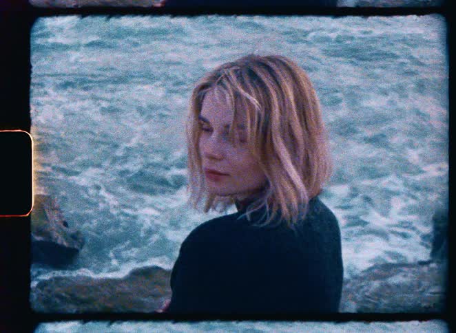 Nostalgic Super 8mm film-style shot of a pensive blonde woman looking at the camera while standing by the choppy ocean