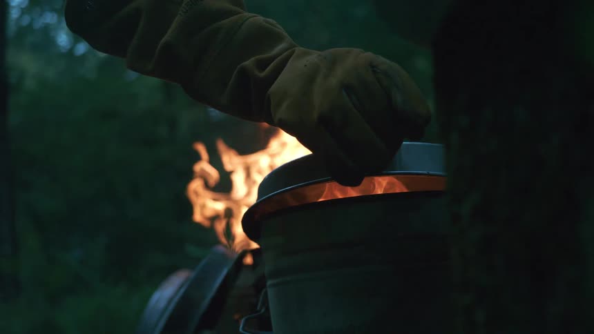 In slow motion, a person wearing a protective glove carefully puts a lid on a fiery metal bucket in a dark forest