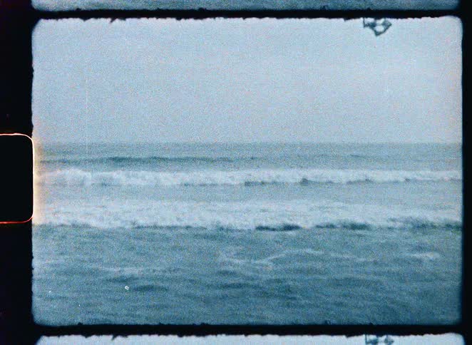 A grainy, vintage 8mm film shot of surfers riding the waves on an overcast day