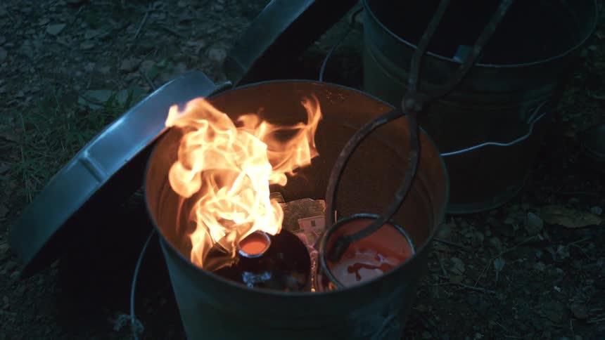 Slow motion shot of Raku pottery being fired in a metal bucket kiln outdoors at dusk, with tongs holding a molten glazed pot