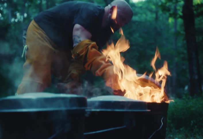 A craftsman in protective gear carefully tends to a fire in metal buckets, creating a cloud of smoke during an outdoor pottery firing