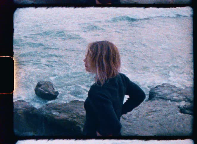 Vintage 8mm footage of a young woman looking back over her shoulder while standing on a rocky coast with crashing waves