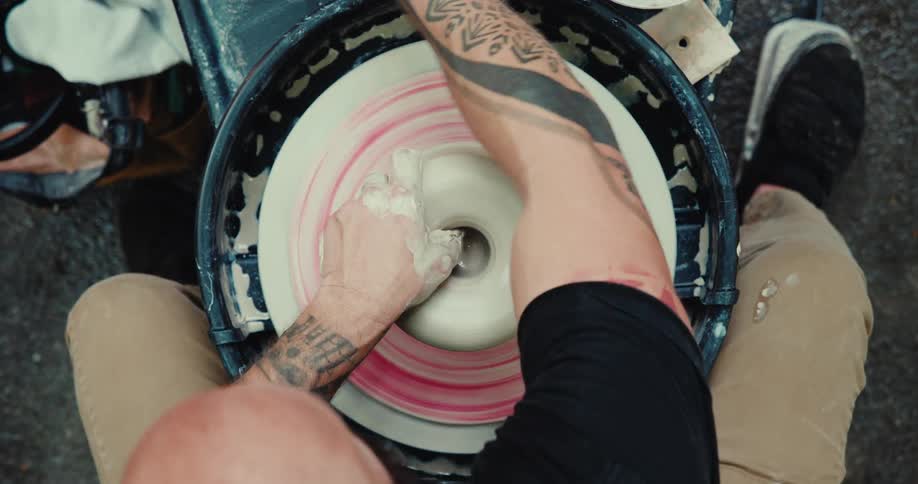 A top-down view shows a tattooed man's hands skillfully molding a piece of clay on a fast-spinning potter's wheel