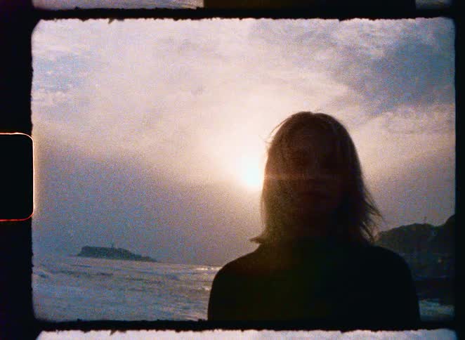 A grainy, vintage film-style shot of a woman in silhouette standing by the ocean at sunset as the light fades