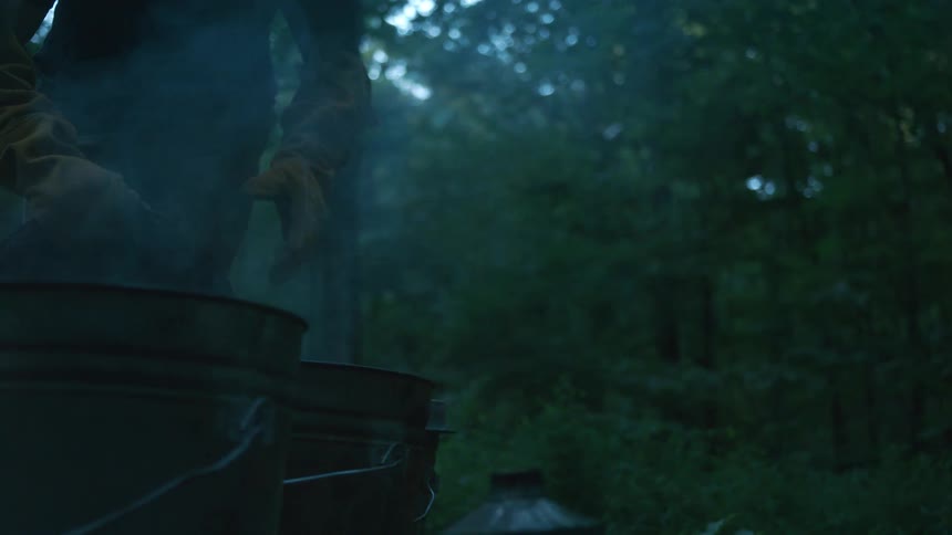 A beekeeper in protective gloves tends to a smoker, with plumes of smoke rising in a dark and moody forest setting