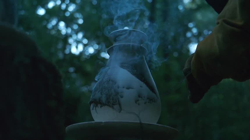 A potter's gloved hand carefully handles a hot, steaming vase during the outdoor Raku firing process