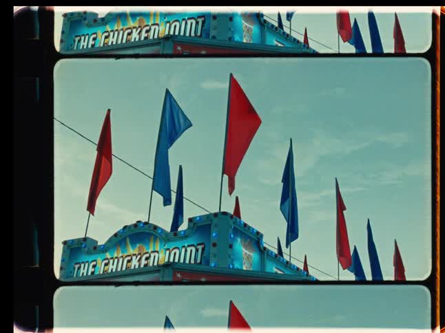 Red and blue flags wave in the wind above a carnival food booth in this retro, film-strip style footage
